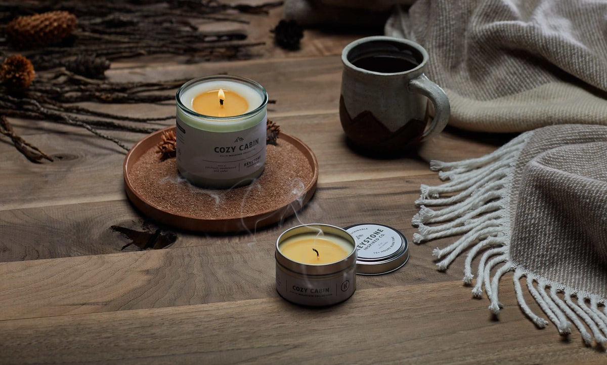 A cozy tabletop scene with two candles from Cozy Candles. One candle is in a clear glass jar with a white label and a lit wick, sitting on a round wooden coaster.