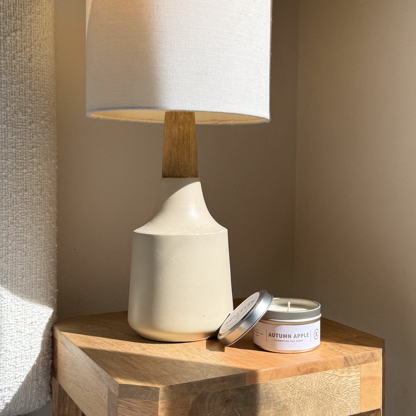 Table lamp with a white shade and wooden base on a wooden surface next to an 'Autumn Apple' candle.
