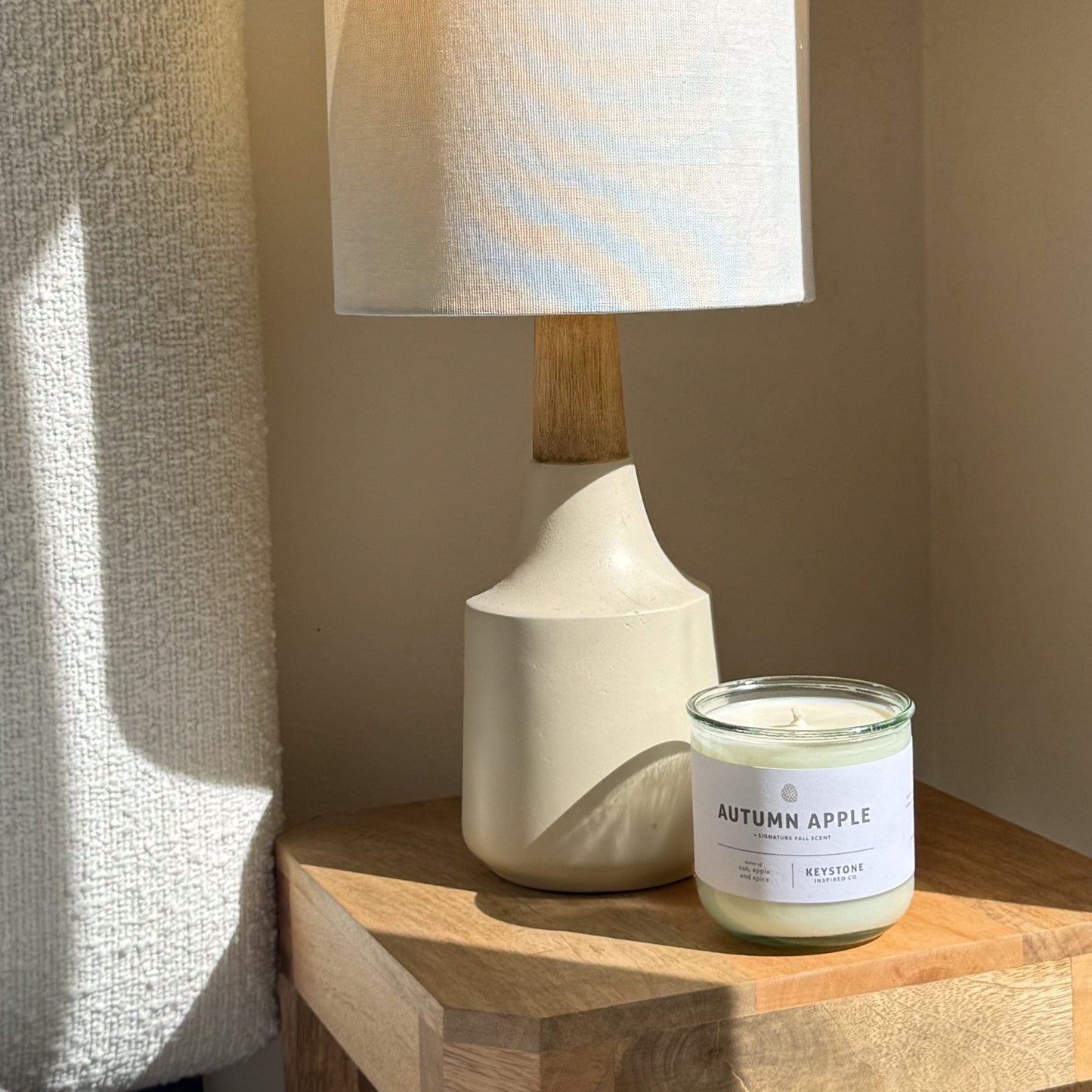 Candle labeled 'Autumn Apple' on a wooden surface next to a lamp.