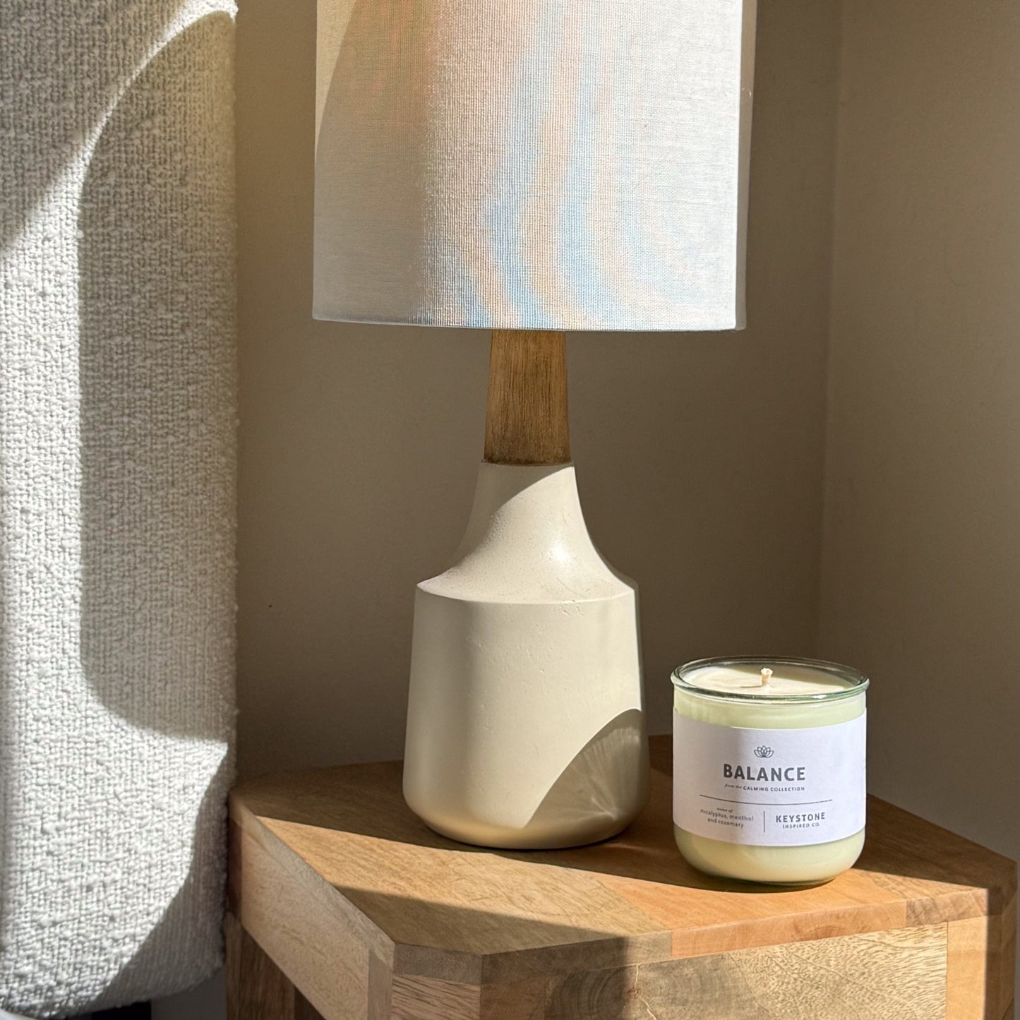 Table lamp with wooden base and white shade next to a 'Balance' candle on a wooden surface.