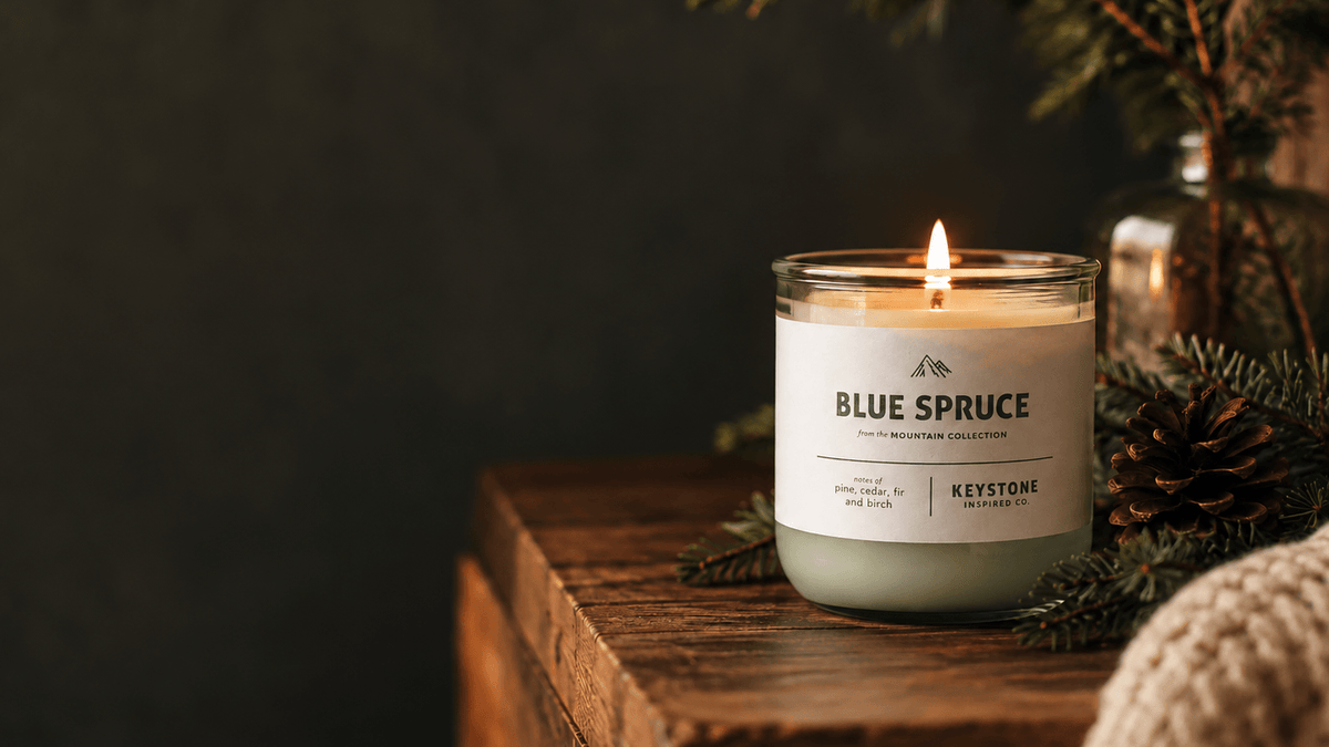 A lit candle in a clear glass jar sits on a rustic wooden surface, surrounded by evergreen branches, a pinecone, and a knitted textile against a dark background. The label reads BLUE SPRUCE and KEYSTONE.