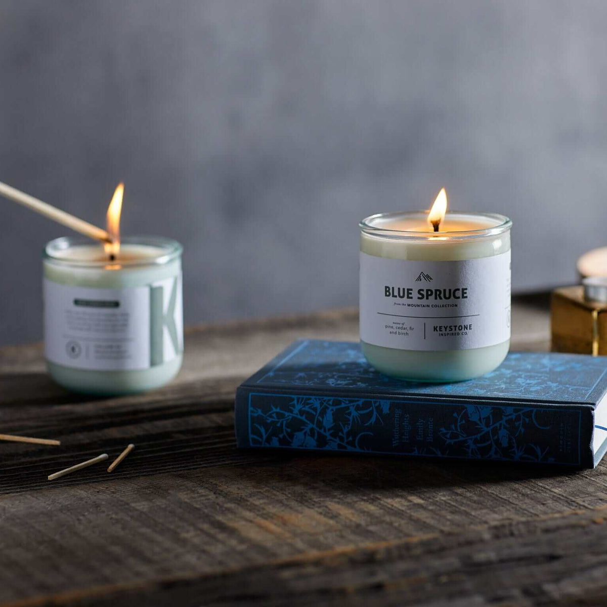 Two lit glass jar candles on a rustic wooden table, with one candle labeled 'BLUE SPRUCE' resting on a blue book and the other being lit with wooden wicks. A small gold container and a wooden dish are in the background against a dark gray wall.
