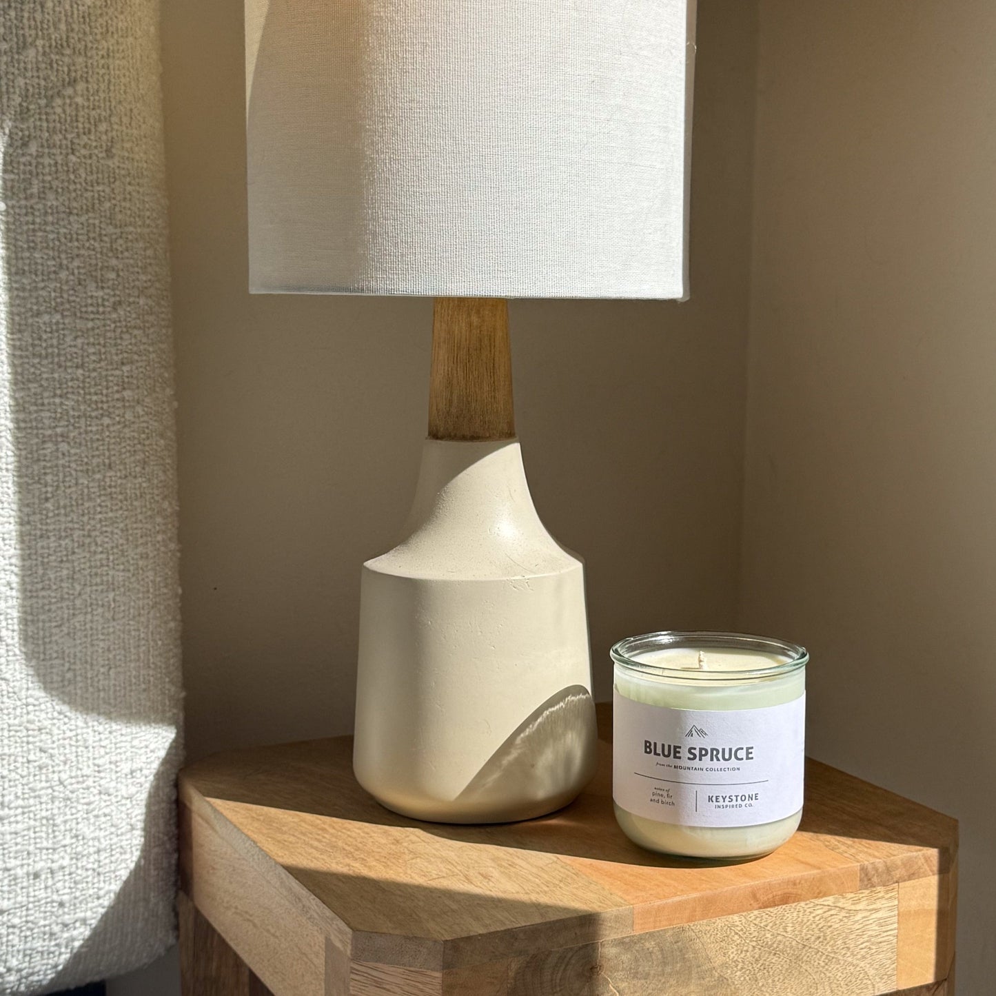White ceramic lamp with wooden base next to a 'Blue Spruce' candle on a wooden surface.