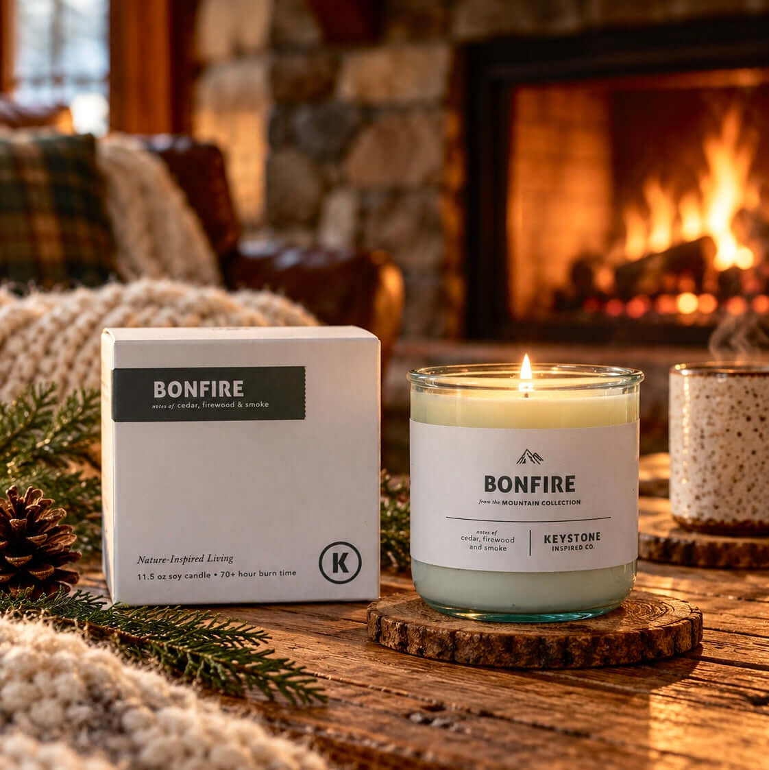 A Bonfire candle in a clear glass jar with a lit flame sits on a rustic wooden coaster beside its white product box on a wooden table in a cozy living room with a fireplace in the background.