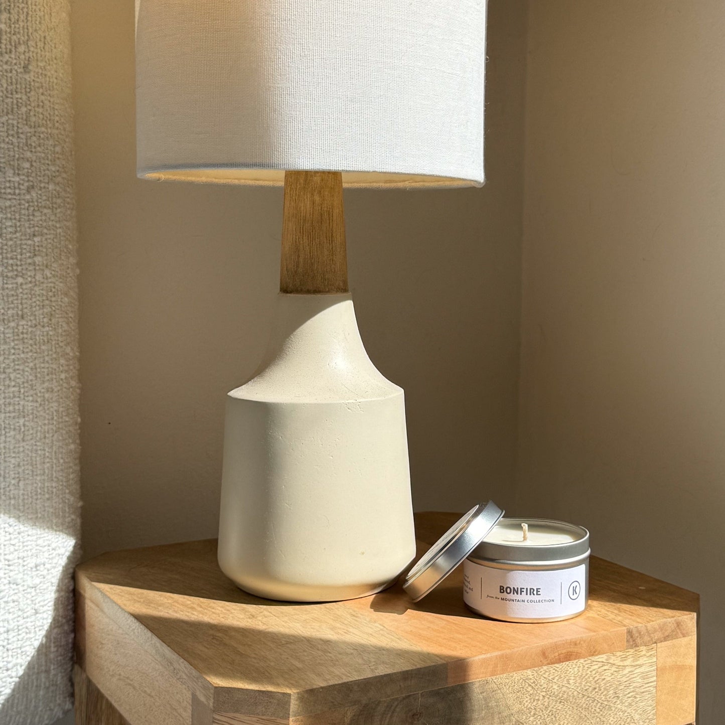 White ceramic lamp with wooden base on a wooden surface next to a 'Bonfire' candle.
