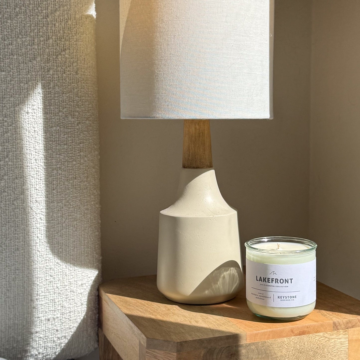 Table lamp with wooden base and white shade next to a candle labeled 'Lakefront' on a wooden surface.