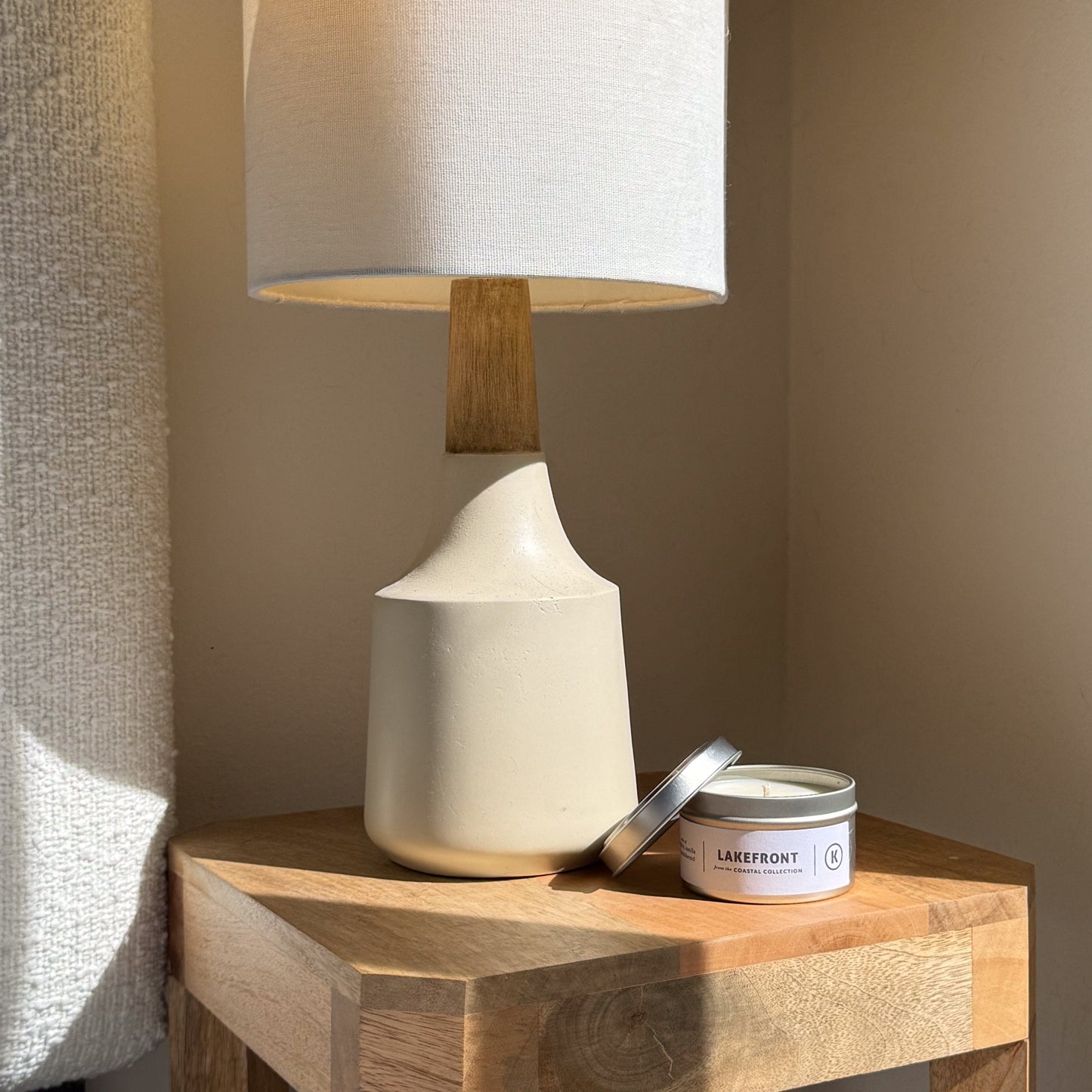 White ceramic lamp with wooden base on a wooden side table next to a candle labeled 'Lakefront'.