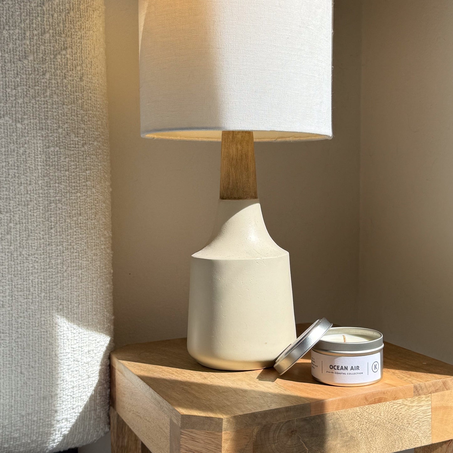 White ceramic lamp with wooden base on a wooden side table next to an 'Ocean Air' candle.
