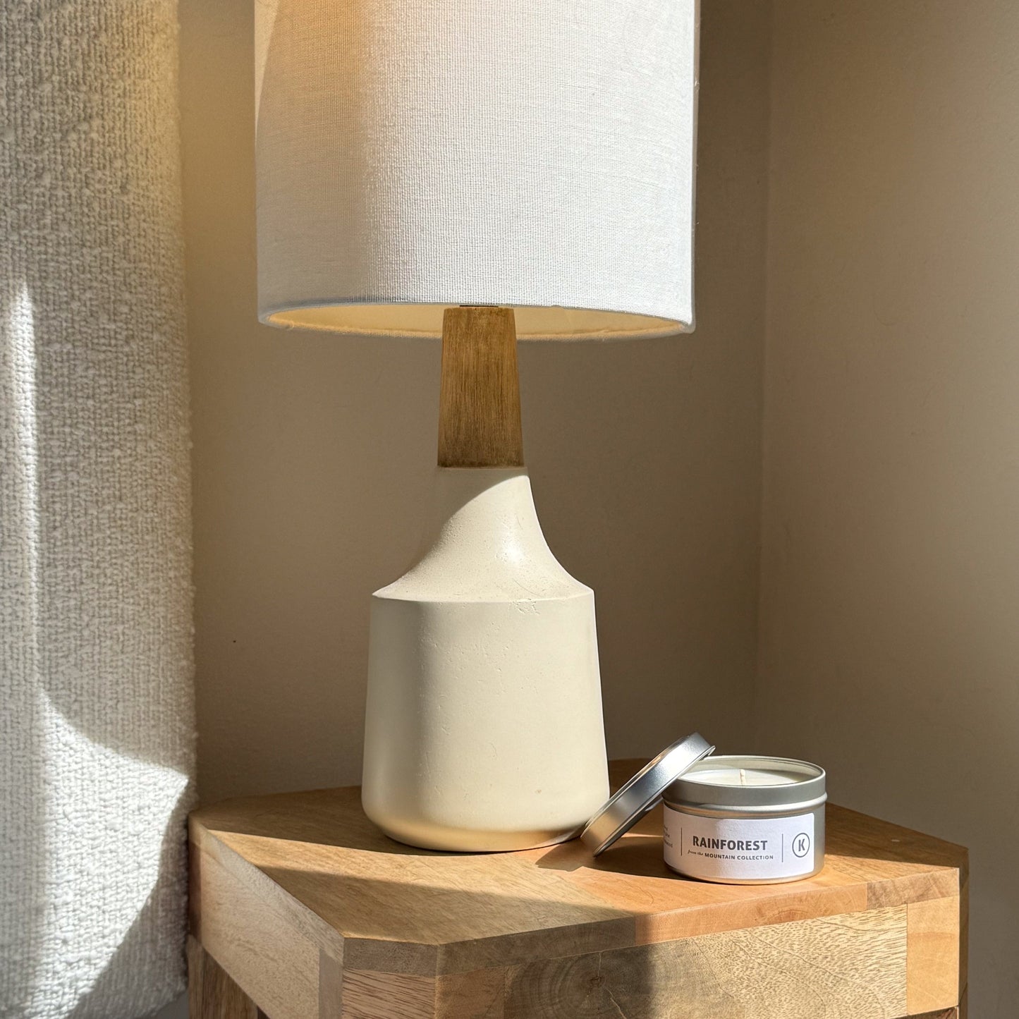 White ceramic lamp with wooden base on a wooden surface next to a candle labeled 'Rainforest'.