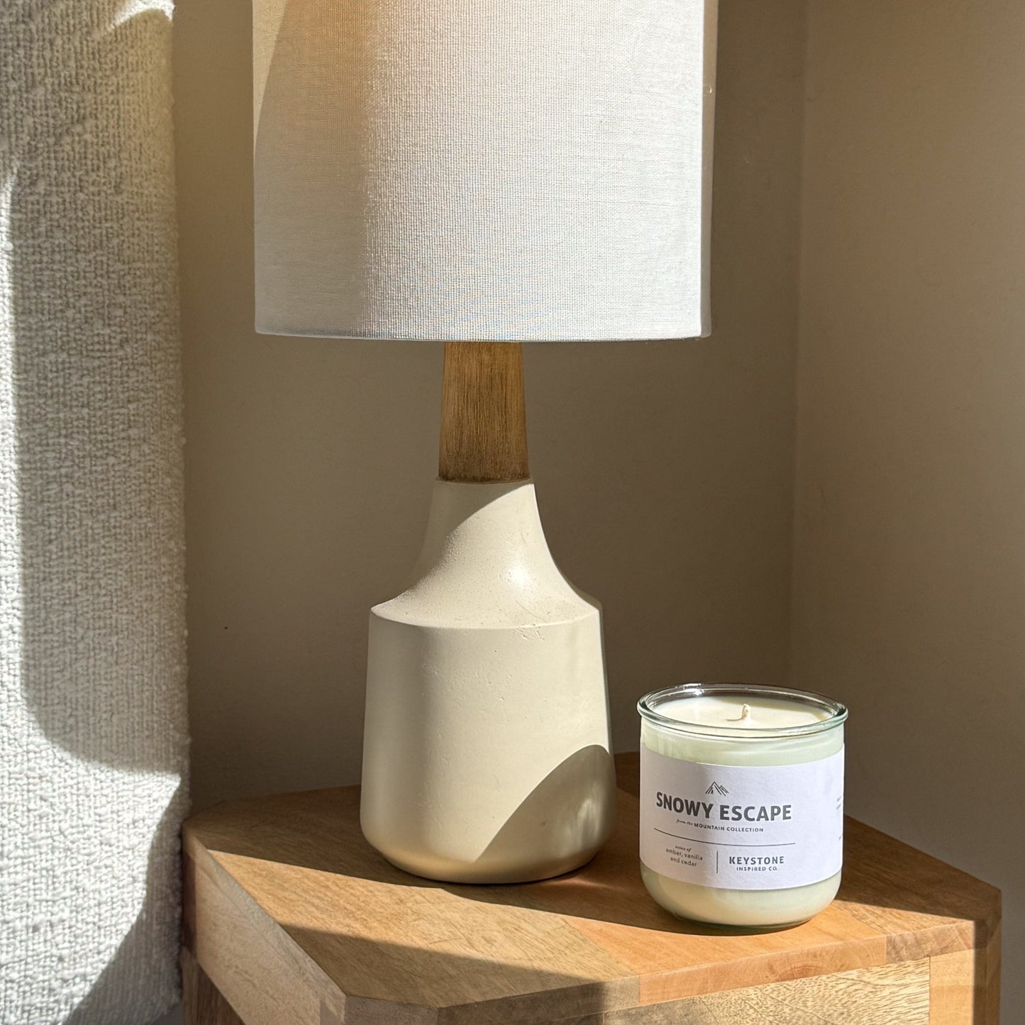 White ceramic lamp with wooden base next to a candle labeled 'Snowy Escape' on a wooden surface.