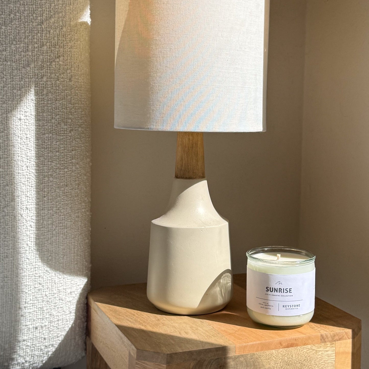 White ceramic lamp with wooden base next to a candle labeled 'Sunrise' on a wooden surface.